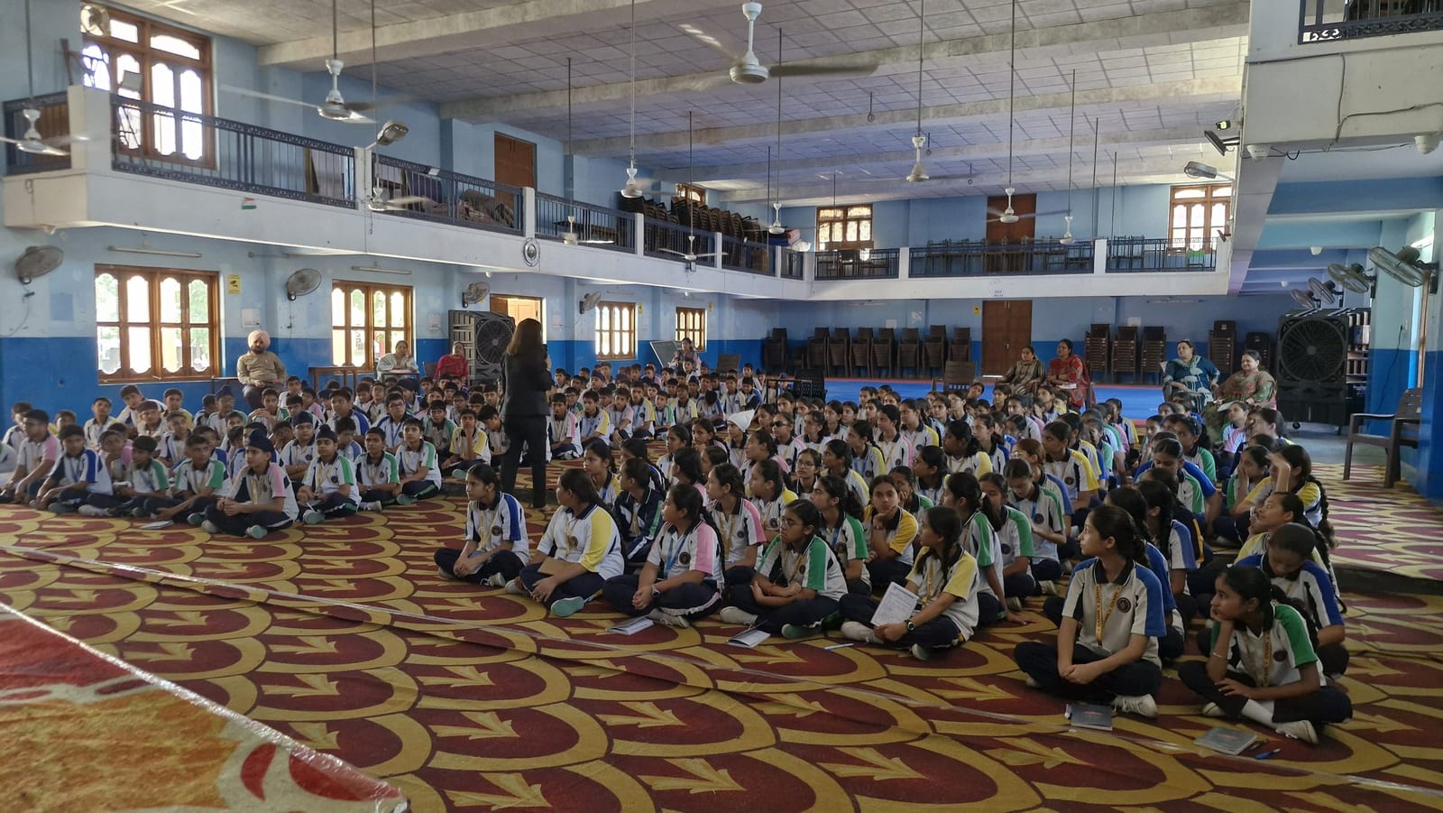 Students seated cross-legged on a patterned carpet, listening to a facilitator standing among them.