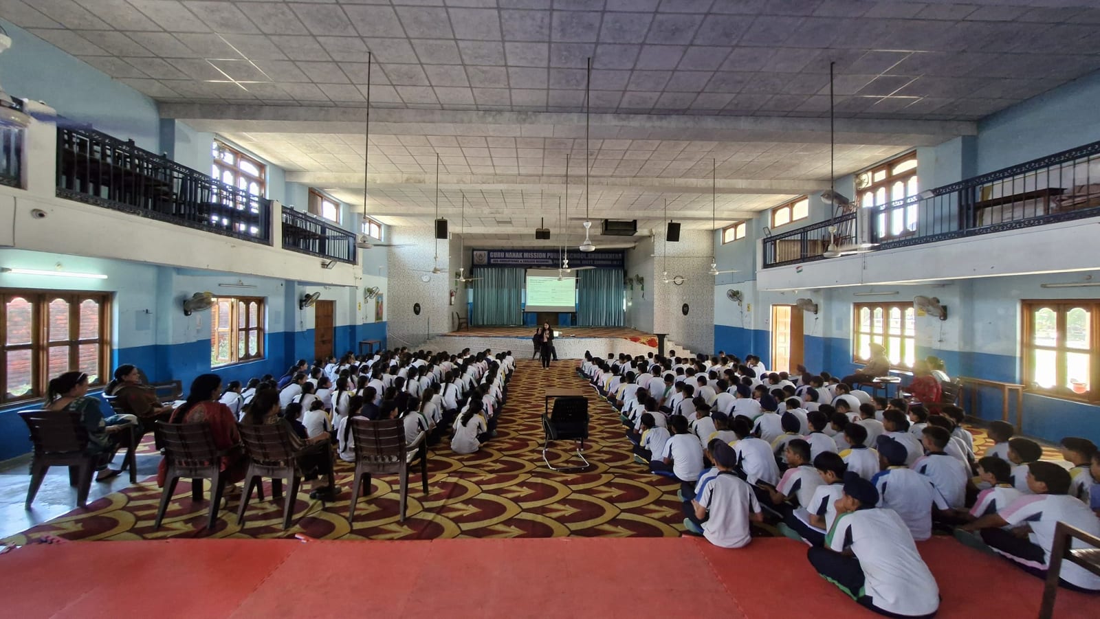A wide view of a school hall mid-session, students seated in rows across the floor.