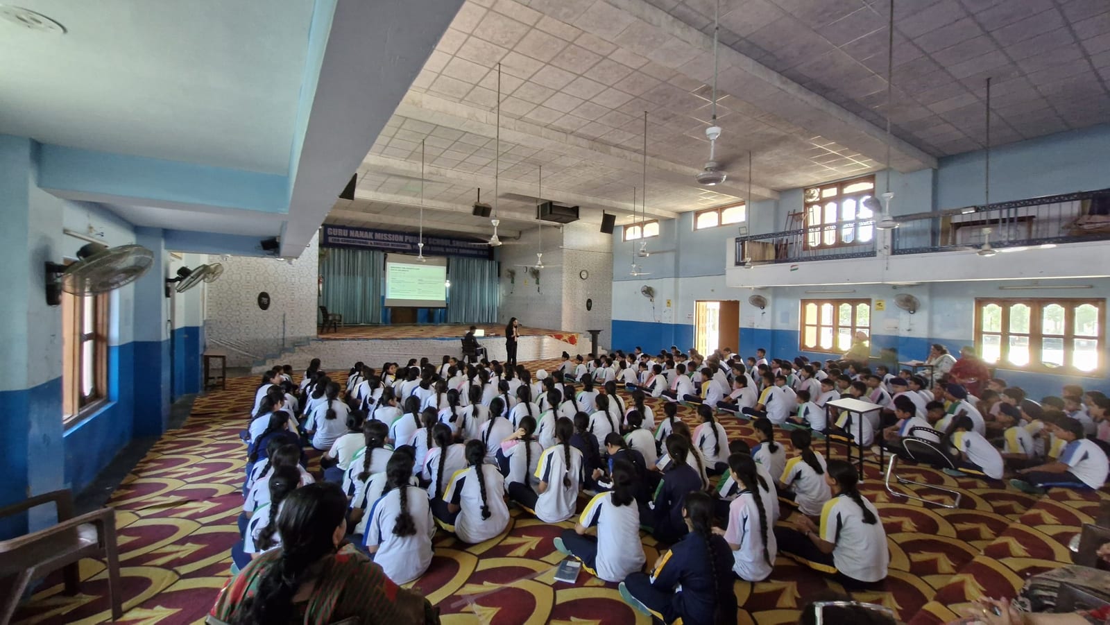 A full school hall seen from the back, students seated on the floor facing a speaker at the front.